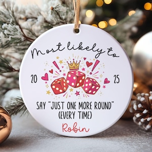 May include: A round white ceramic ornament hanging from a gold ribbon. The design features three red dice, a small gold crown, pink hearts, and stars. Text reads: Most likely to 20 25 SAY "JUST ONE MORE ROUND" (EVERY TIME) and the name Robin in red.