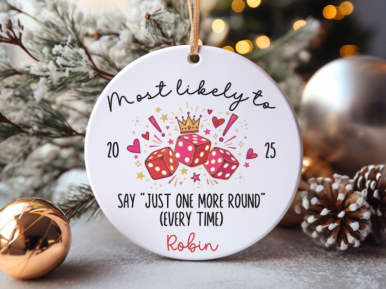 May include: A round white ceramic ornament hanging from a gold ribbon. The design features three red dice, a small gold crown, pink hearts, and stars. Text reads: Most likely to 20 25 SAY "JUST ONE MORE ROUND" (EVERY TIME) and the name Robin in red.
