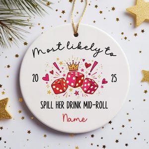 May include: A round white ceramic ornament with a gold hanging string. The design features three red dice, a gold crown, pink hearts, and stars. Text reads: Most likely to 20 25 SPILL HER DRINK MID-ROLL and the word Name in red script.