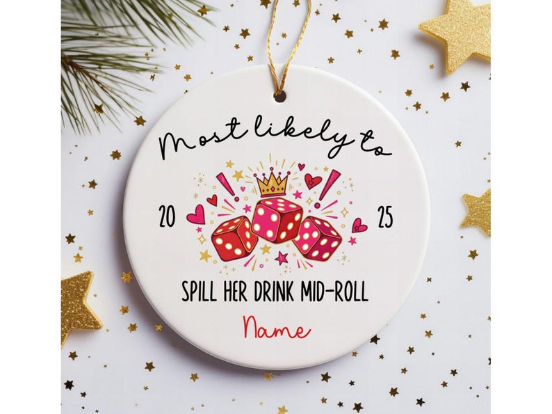 May include: A round white ceramic ornament with a gold hanging string. The design features three red dice, a gold crown, pink hearts, and stars. Text reads: Most likely to 20 25 SPILL HER DRINK MID-ROLL and the word Name in red script.