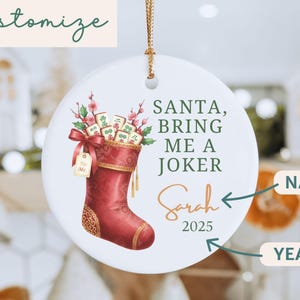 May include: A round white ceramic ornament featuring a red Christmas stocking filled with Mahjong tiles and pink flowers. The text reads SANTA, BRING ME A JOKER, Sarah, and 2025. The ornament hangs from a gold string and includes a gift tag.