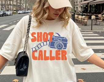 May include: An off-white short-sleeve t-shirt featuring a graphic print of a blue camera with a film strip and the orange text SHOT CALLER. The shirt is paired with a black quilted shoulder bag with a gold chain strap and a beige baseball cap.