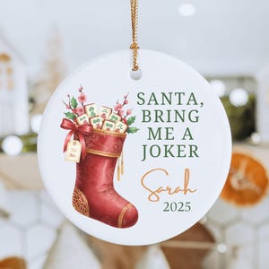 May include: A round white ceramic ornament featuring a red Christmas stocking filled with mahjong tiles and pink floral sprigs. The text reads SANTA, BRING ME A JOKER, Sarah, and 2025. A gold string is attached to the top for hanging purposes.