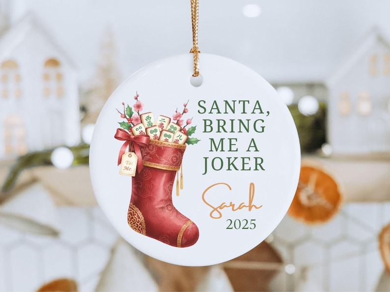 May include: A round white ceramic ornament featuring a red Christmas stocking filled with mahjong tiles and pink floral sprigs. The text reads SANTA, BRING ME A JOKER, Sarah, and 2025. A gold string is attached to the top for hanging purposes.