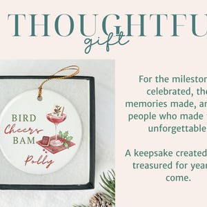 May include: A round white ceramic ornament with a gold hanging string, presented in a black box. The ornament features a graphic of a cocktail glass, a small book, and holly, with the text BIRD CHEERS BAM and the name Polly printed on it.