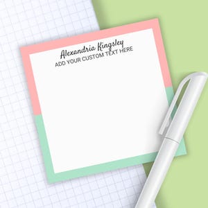 May include: A square notepad with a white center, a pink top border, and a mint green bottom border. The text "Alexandria Kingsley" and "ADD YOUR CUSTOM TEXT HERE" is printed in black at the top. A white pen is positioned next to the notepad.
