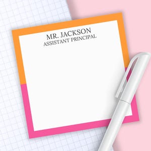 May include: A square notepad with a white center and a border split between orange on the top and pink on the bottom. The text MR. JACKSON ASSISTANT PRINCIPAL is printed in black at the top. A white pen is positioned diagonally across the notepad.