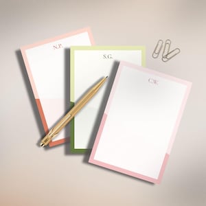 May include: Three personalized notepads with pastel borders in pink, green, and coral, each with initials. A gold pen and three gold paper clips are also present. The notepads are arranged on a light-colored surface.