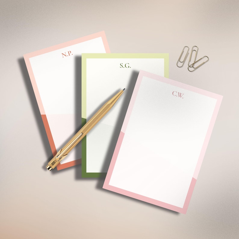 May include: Three personalized notepads with pastel borders in pink, green, and coral, each with initials. A gold pen and three gold paper clips are also present. The notepads are arranged on a light-colored surface.