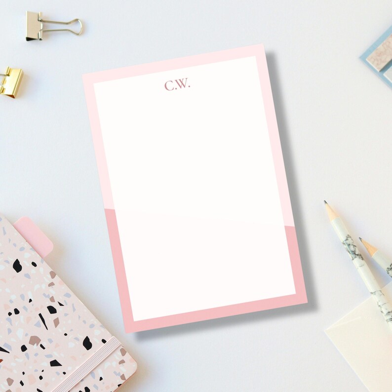 May include: A pink and white notepad with the initials "C.W." in the upper left corner. The notepad has a pink border and a pink accent at the bottom. The notepad is on a white surface with office supplies.