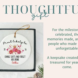 May include: A round white ceramic ornament with a gold hanging string, presented in a black box. The ornament features three red dice, a gold crown, and the text: Most likely to 20 16 CHANGE SEATS AND FORGET HER DRINK Dottie, surrounded by small hearts.