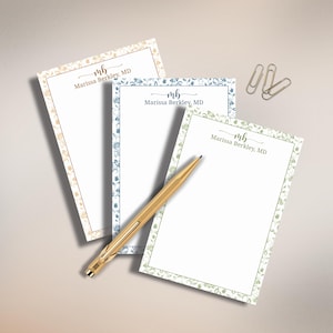 May include: Three rectangular stationery notepads with floral borders in peach, blue, and green. Each notepad features the monogram mb and the text Marissa Berkley, MD. A gold-toned metal pen rests diagonally across the notepads, and two silver paper clips are placed near the top right corner.
