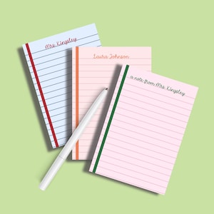 May include: Three lined notepads with different colored binding and text. One notepad is blue with red binding and the text "Mrs. Kingsley". Another is pink with orange binding and the text "Laura Johnson". The third is pink with green binding and the text "a note from Mrs. Kingsley". A white pen is in the foreground.