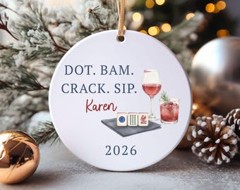 May include: A round white ceramic ornament featuring the text DOT. BAM. CRACK. SIP. Karen 2026 in blue and red. The design includes illustrations of three mahjong tiles on a grey napkin, a glass of red wine, and a cocktail with a sprig of rosemary.