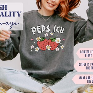 May include: A dark gray sweatshirt with the text "PEDS ICU" in white letters. The text is surrounded by two red strawberries with green leaves and white flowers. The sweatshirt has a relaxed fit and is made of a medium-heavy fabric.