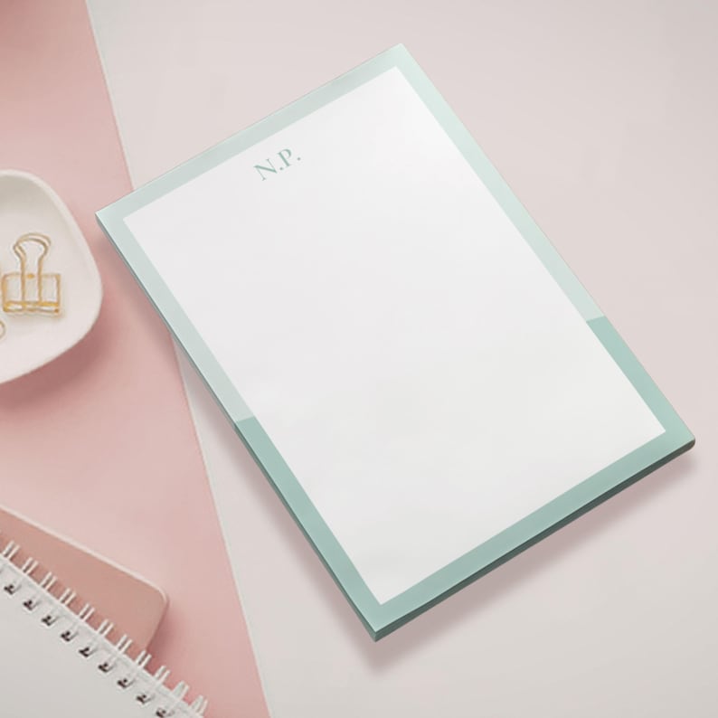 May include: A rectangular notepad with a white interior and a light green border. The initials "N.P." are printed in the upper left corner. The notepad is on a pink and white surface, with a small dish and gold paper clip visible.