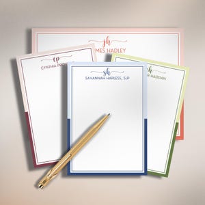 May include: A collection of four personalized stationery notepads in various colors including light blue, pink, and green, each featuring a monogram and a name printed at the top. A gold-toned metal pen rests diagonally across the front notepad.