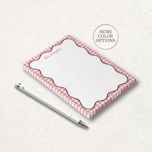 May include: A personalized notepad with a white writing surface and a red plaid border. The notepad has a scalloped edge and the name "Mrs. Kingsley" printed in the upper left corner. A silver pen rests beside the notepad. The image also includes a circular graphic that says "MORE COLOR OPTIONS".
