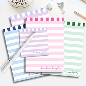 May include: A collection of colorful striped notepads in various sizes. The pads feature horizontal stripes in shades of blue, pink, purple, and green, with a solid color border at the top. Some pads have a name printed at the bottom.