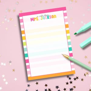 May include: A notepad with a pink, orange, and white striped border and a colorful striped interior. The text "a note from Mrs. Jackson" is at the top. A green pen and two green markers are on the pink background.