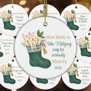 May include: A round white ceramic ornament featuring a green Christmas stocking filled with Mahjong tiles, holly, and berries. The text reads Most likely to Take Mahjong way too seriously, Sherry, and 2026, with a gold hanging string at the top.