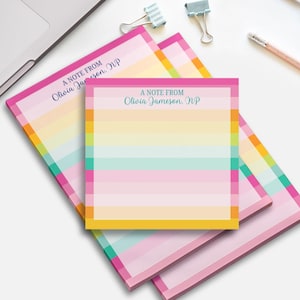 May include: A stack of colorful notepads with a rainbow striped design. Each notepad has a pink border and the text "A NOTE FROM Olivia Jameson, NP" in a light blue font. Pencils and paper clips are visible in the background.