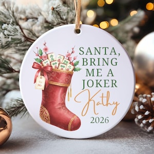 May include: A round white ceramic ornament featuring a red Christmas stocking filled with Mahjong tiles and pink floral sprigs. The text reads: SANTA, BRING ME A JOKER, Kathy, 2026. A small gift tag attached to the stocking says To: Me.