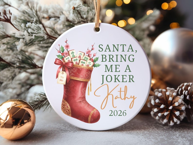 May include: A round white ceramic ornament featuring a red Christmas stocking filled with Mahjong tiles and pink floral sprigs. The text reads: SANTA, BRING ME A JOKER, Kathy, 2026. A small gift tag attached to the stocking says To: Me.