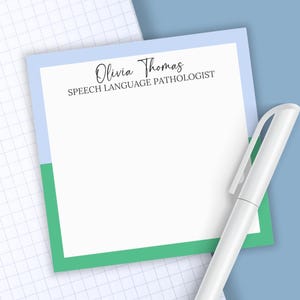 May include: A square notepad with a white center and a border featuring light blue on the top and green on the bottom. The text Olivia Thomas SPEECH LANGUAGE PATHOLOGIST is printed in black at the top. A white pen is positioned next to the notepad.