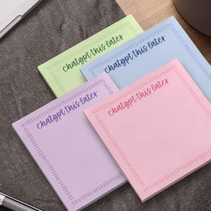 May include: A stack of square sticky notes in pastel colors: green, blue, purple, and pink. Each note has the phrase "chatgpt this later" in a handwritten style font. A pen and laptop are in the background.