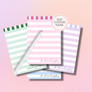 May include: A collection of personalized notepads with horizontal stripes in various pastel colors: pink, green, purple, and blue. Each notepad features a custom name and a white pen rests across the pads. The design includes the text "ADD CUSTOM NAME".