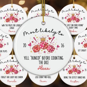 May include: A round white ceramic ornament featuring a graphic of three pink dice, a gold crown, and red hearts. The text reads "Most likely to YELL "BUNCO!" BEFORE COUNTING THE DICE" with the name "Alexa" and the years "20" and "26" printed in black and red.