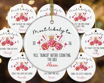May include: A round white ceramic ornament featuring a graphic of three pink dice, a gold crown, and red hearts. The text reads "Most likely to YELL "BUNCO!" BEFORE COUNTING THE DICE" with the name "Alexa" and the years "20" and "26" printed in black and red.