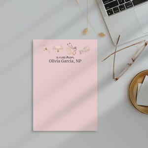 May include: A pink notepad with a note from Olivia Garcia, NP. The notepad has a watercolor illustration of medical supplies, including a stethoscope, a pill bottle, a syringe, and a medical bag.