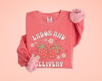 Op de afbeelding: Een roze sweatshirt met de tekst "Arbeid en Bevalling" in een retro-lettertype, met twee aardbeien en bloemen die op de voorkant zijn gedrukt.