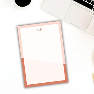 May include: A personalized notepad with a two-tone design. The top and sides are a light pink, while the bottom is a darker coral color. The initials "N.P." are printed in a matching coral color at the top.