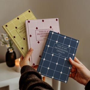 May include: Three hardcover notebooks in yellow, light pink, and dark blue, each featuring a white grid pattern with diamond accents and a central rectangular box containing the text THIS IS MY PROFESSIONAL OPINION and the name Oliver.
