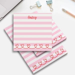 May include: Pink and white striped notepad with the name "Haley" in red script. The bottom edge features a red heart design. Several notepads are stacked on a white surface, with office supplies in the background.
