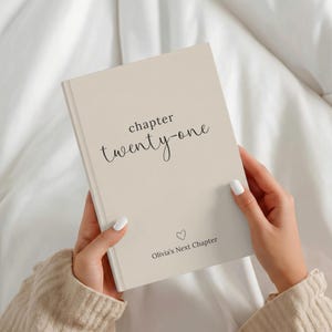 May include: A beige hardcover book with black text that reads chapter twenty-one and Olivia's Next Chapter, featuring a small heart icon centered below the text. The book has a smooth, matte finish and is held by two hands.