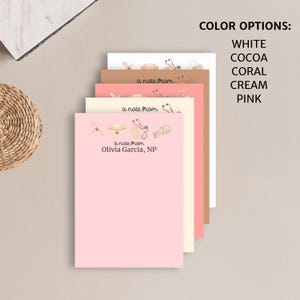 May include: A stack of pink, cream, coral, cocoa, and white notepads with a medical theme. The top notepad has the text "a note from Olivia Garcia, NP" and features a stethoscope, bandages, a nurse's hat, and other medical supplies. The text "COLOR OPTIONS: WHITE COCOA CORAL CREAM PINK" is visible on the right side of the image.