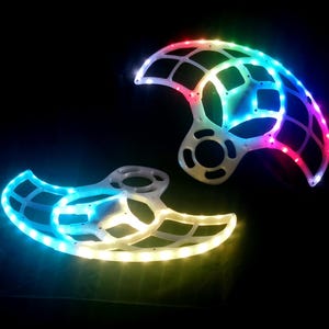 Pode incluir: Duas estruturas geométricas brancas e iluminadas com luzes LED coloridas. As luzes percorrem um espectro de cores, incluindo azul, verde e vermelho, sobre um fundo preto. As estruturas têm um design semelhante a uma grelha.