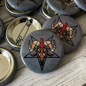 Puede incluir: Una colección de botones redondos grises con un pentagrama y una cabeza de cabra con una cruz roja sobre ella. Los botones tienen un respaldo plateado con un pasador de seguridad.