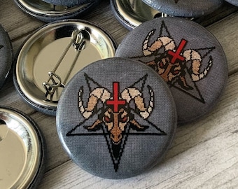 Könnte beinhalten: Eine Sammlung runder, grauer Knöpfe mit einem Pentagramm und einem Ziegenkopf mit einem roten Kreuz darüber. Die Knöpfe haben eine silberne Rückseite mit einer Sicherheitsnadel.
