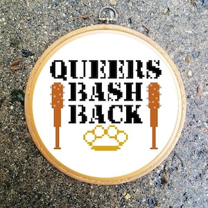 Könnte beinhalten: Weißer Stickrahmen mit dem Text "QUEENS BASH BACK" in schwarzer Pixel-Schrift. Zwei braune pixelartige Schläger befinden sich auf beiden Seiten des Textes. Ein goldener pixelartiger Schlagring befindet sich unter dem Text.