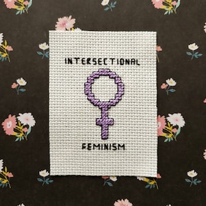 May include: A white cross-stitch with a purple female symbol and the text "INTERSECTIONAL FEMINISM".