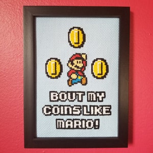 May include: A framed cross-stitch design featuring a pixelated image of Mario from the Super Mario Bros. video game collecting coins. The text below reads "Bout my coins like Mario!"