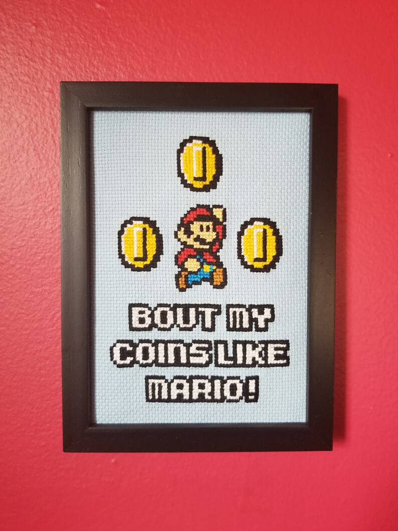 May include: A framed cross-stitch design featuring a pixelated image of Mario from the Super Mario Bros. video game collecting coins. The text below reads "Bout my coins like Mario!"