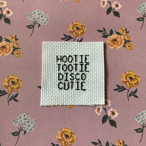 Puede incluir: Un punto de cruz blanco con bordado negro que dice "Hootie Tootie Disco Cutie" sobre un fondo floral rosa.