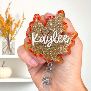 Puede incluir: Un portatarjetas con forma de hoja de arce con purpurina dorada y detalles en naranja y rojo. El nombre "Kaylee" est&aacute; escrito en blanco. El portatarjetas tiene un cord&oacute;n retr&aacute;ctil de pl&aacute;stico transparente.