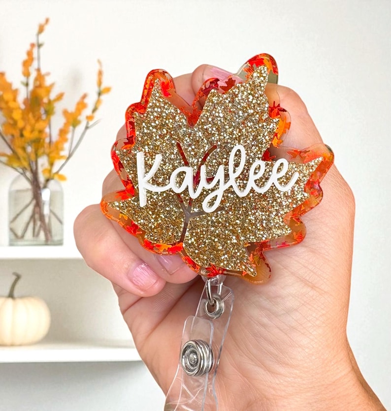 Puede incluir: Un portatarjetas con forma de hoja de arce con purpurina dorada y detalles en naranja y rojo. El nombre "Kaylee" est&aacute; escrito en blanco. El portatarjetas tiene un cord&oacute;n retr&aacute;ctil de pl&aacute;stico transparente.